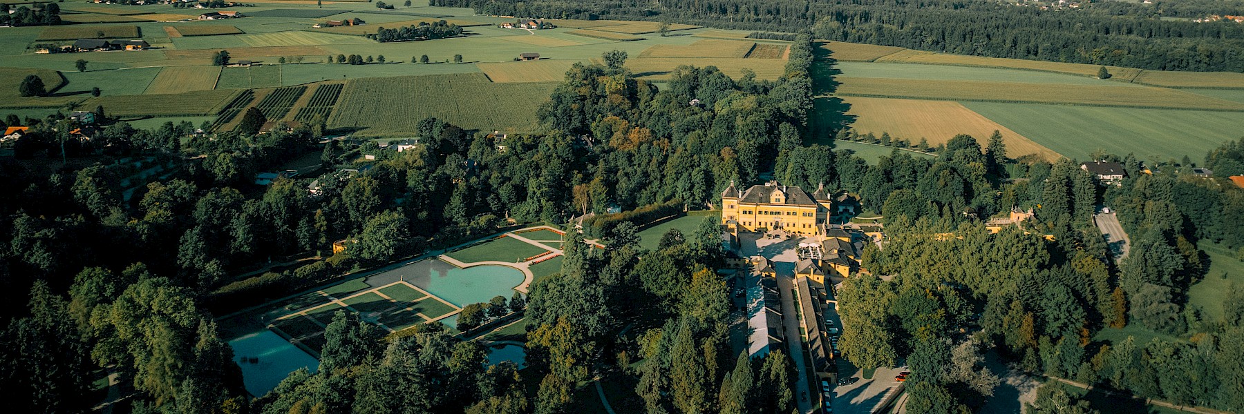 Heiraten in Hellbrunn (c) Gassner Gastronomie, Fotografie Voglperspektive: Diese liebevoll gestaltete Kulisse für eine Trauung im Freien befindet sich inmitten eines weitläufigen Parks, umgeben von hohen, grünen Bäumen. Ein weißer Teppich führt durch zwei symmetrische Reihen eleganter, weißer Stühle, die mit zarten Blumengestecken geschmückt sind. Große, weiße Luftballons, die an den Stühlen befestigt sind, verleihen der Szenerie eine verspielte Leichtigkeit. Am Ende des Weges steht ein schlichter, stilvoll dekorierter Trautisch, eingerahmt von Blumen. Die Atmosphäre ist ruhig, romantisch und perfekt für eine unvergessliche Hochzeit inmitten der Natur.