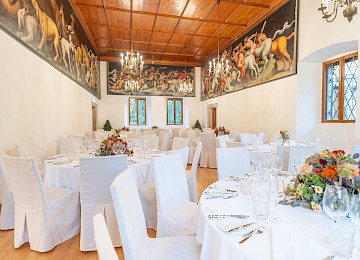 The image shows a magnificent banquet hall set for an elegant celebration. The high wooden ceiling and dramatic wall murals depicting mythical and animal scenes lend the room a grand, historic flair. White-covered chairs and round tables are arranged neatly, adorned with vibrant floral centerpieces in red, pink, and orange hues. Tall candelabras add a touch of romantic charm. Natural light streams through the windows, enhancing the warm tones of the decor. This impressive setting blends classic art and refined festivity—ideal for a wedding or formal event in a castle-like atmosphere.
